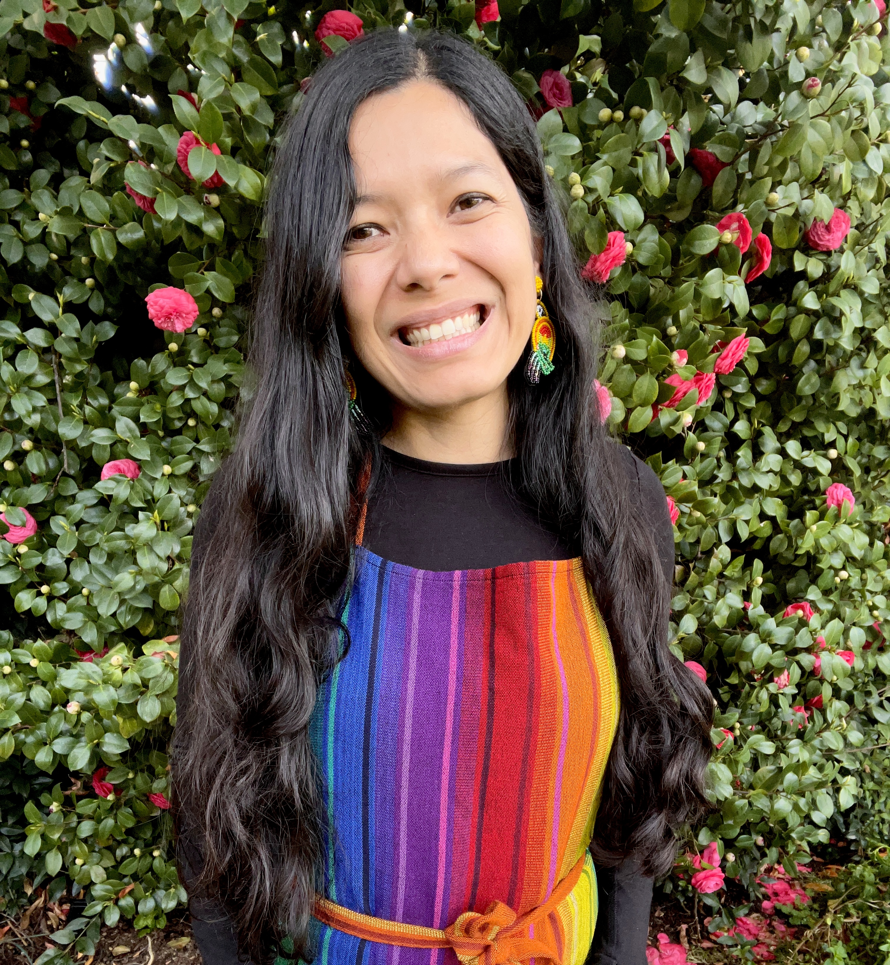 Photo of Toni Okamoto standing in front of a camelia bush and wearing a rainbow apron.