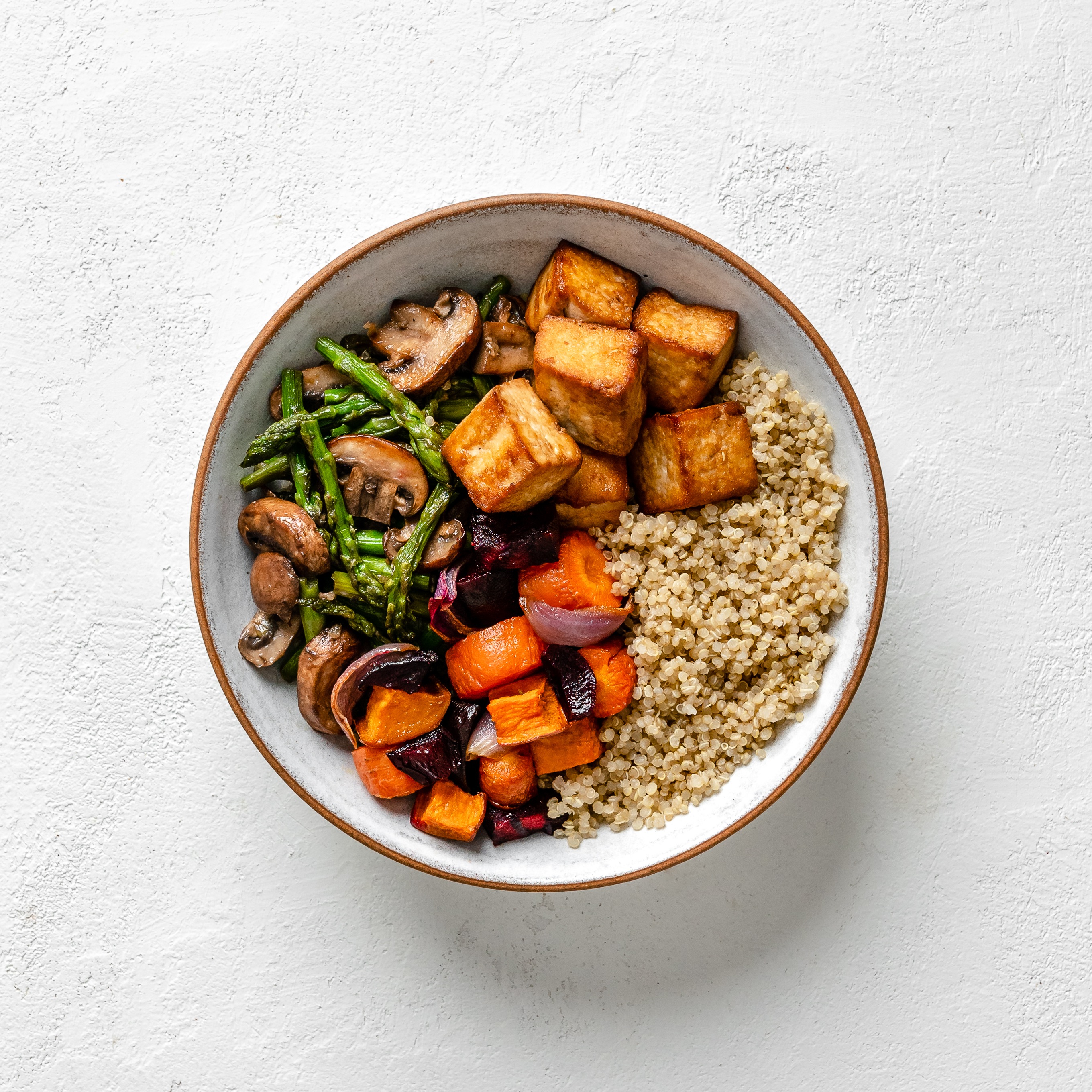 White bowl filled with tofu, vegetables, and quinoa on a white textured background